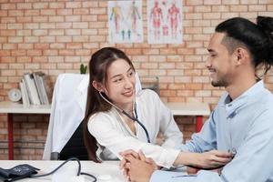 Hermosa mujer médico con camisa blanca que es una persona asiática con estetoscopio es salud examinando a un paciente masculino en la clínica médica de fondo de pared de ladrillo, sonriendo asesorando a la ocupación del especialista médico. foto