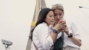 Asian lesbian couples using smartphones browsing the website while standing on the bridge. video
