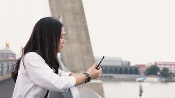 mujer asiática que usa el teléfono inteligente charlando con amigos y navegando por las redes sociales en un teléfono móvil mientras está de pie en el puente. video