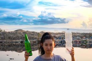 A woman stands hand holding green glass bottle and clear plastic bottle at mountain large garbage background photo
