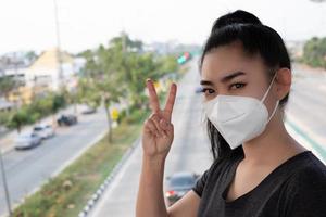 Woman standing hand sign of 2 fingers with putting on respirator N95 mask to protect from airborne respiratory diseases as the flu covid-19 coronavirus PM2.5 dust and smog on the road burred backgro photo