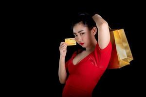 Portrait of a beautiful young Asia woman wearing a red dress holding credit card and shopping bags over a black background, sale concept, look at the camera photo