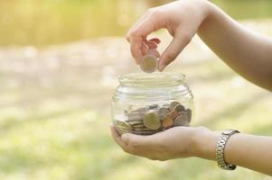 women's hand put money coins in glass bottle for save and donation concept on green garden background