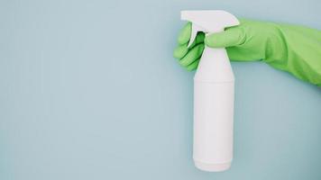 Cerrar la mano del limpiador con guantes verdes sosteniendo una botella de spray blanco como telón de fondo azul