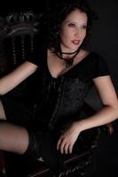 Beautiful green eyed young woman in black corset with stone necklace sitting facing side. photo