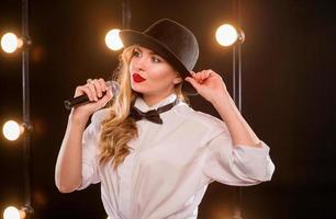 Joven mujer atractiva rubia con camisa blanca, mariposa, sombrero negro con micrófono cantando en el escenario foto