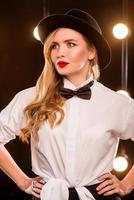 Joven mujer atractiva rubia con camisa blanca, mariposa, sombrero negro con micrófono cantando en el escenario