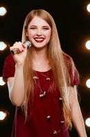 Sonriente joven hermoso cabello largo en vestido rojo mujer con micrófono cantando canciones en el escenario de karaoke foto