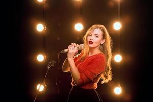 Sonriente joven hermoso cabello largo en suéter rojo niña con micrófono cantando canciones en el escenario de karaoke