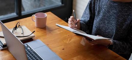 Cropped photo hand of woman writing making list taking notes in notepad working or learning online with laptop at home