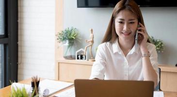 Sonriente mujer asiática que toma una llamada en el teléfono móvil durante el trabajo en una computadora portátil para obtener el número de contabilidad financiera, informe mensual de estadísticas de trámites