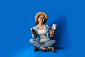 chica viajera con un vestido azul está sonriendo feliz con una cara alegre y sosteniendo un pasaporte con teléfonos móviles en la mano, sobre fondo de color azul. concepto de viaje foto