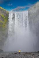 Powerful and famous Skogafoss waterfall with a lonely standing person in orange jacket, while hiking in Iceland, summer, scenic dramatic view at sunny day and blue sky. photo