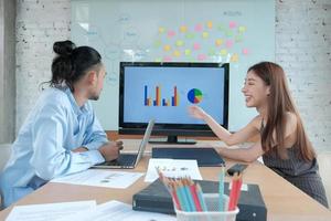 Two young and smart Asian co-workers are brainstorming at office. Female colleague presents business chart to male partner using a laptop in a meeting room, discussing the company's finances project. photo