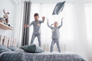 Happy kids playing in white bedroom. Little boy and girl, brother and sister play on the bed wearing pajamas. Nightwear and bedding for baby and toddler. Family at home photo