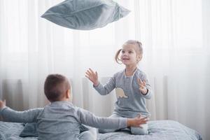Happy kids playing in white bedroom. Little boy and girl, brother and sister play on the bed wearing pajamas. Nightwear and bedding for baby and toddler. Family at home photo