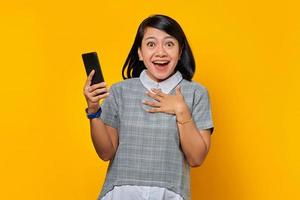 Cheerful young Asian woman holding mobile phone and putting palms on chest over yellow background photo