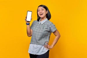 Alegre mujer asiática mostrando la pantalla en blanco del smartphone sobre fondo amarillo