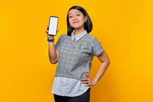 Smiling asian woman showing blank smartphone screen and pointing on it while looking at the camera photo