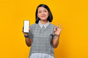 Smiling Asian woman showing smartphone blank screen and gesturing okay sign over yellow background photo