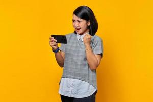 Retrato de mujer asiática joven alegre jugando videojuegos en el móvil y celebrando el éxito sobre fondo amarillo