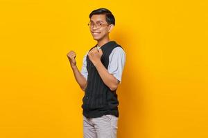 Portrait of excited young Asian man celebrating success with raised arms on yellow background photo