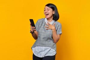 Cheerful young Asian woman holding mobile phone with open mouth on yellow background