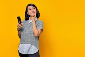 Retrato de mujer asiática joven alegre mirando a un lado pensando en pregunta con el dedo en la barbilla y sosteniendo el teléfono móvil foto