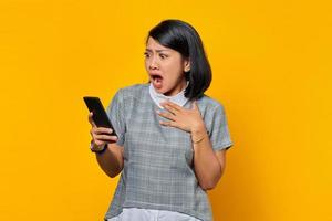 Mujer asiática joven sorprendida que sostiene el teléfono móvil con la boca abierta sobre fondo amarillo
