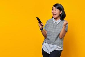 Portrait of excited young Asian woman holding smartphone and celebrating success over yellow background