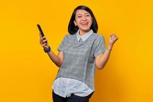 Young Asian woman feeling excited and happy while looking at messages on smartphone on yellow background photo