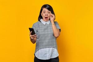 Retrato de conmocionada y sorprendida joven mujer asiática sosteniendo smartphone y gafas mirando a la cámara sobre fondo amarillo foto