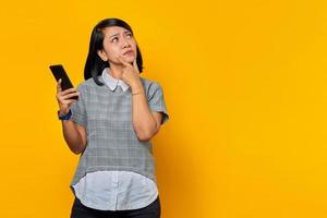 Pensive young Asian woman looking up with finger on chin and holding cell phone over yellow background photo