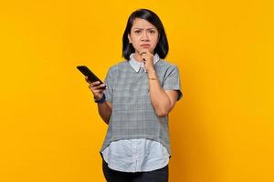 Pensive young Asian woman using mobile phone with fingers on chin over yellow background photo