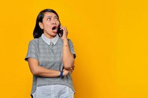 Portrait of shocked young Asian woman while receiving incoming call on smartphone on yellow background photo