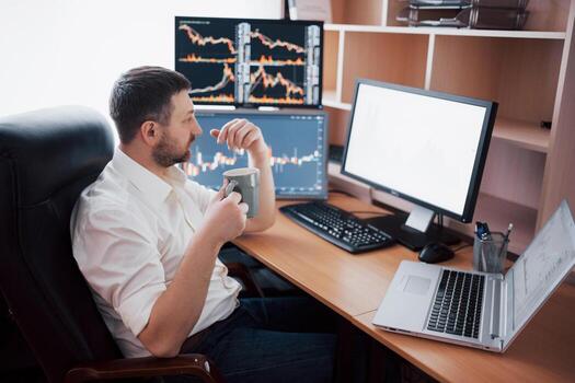 Young businessman is sitting in office at table, working on computer with many monitors,diagrams on monitor. Stock broker analyzes binary options charts.Hipster man drinking coffee,studying photo