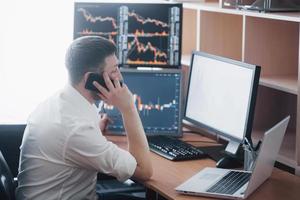 Over the shoulder view of and stock broker trading online while accepting orders by phone. Multiple computer screens ful of charts and data analyses in background photo