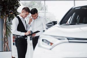 Two men stand in the showroom against cars. Close-up of a sales manager in a suit that sells a car to a customer. The seller gives the key to the customer. photo