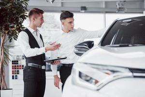 dos hombres se paran en la sala de exposición contra los coches. primer plano de un gerente de ventas con un traje que vende un automóvil a un cliente. el vendedor le da la llave al cliente.