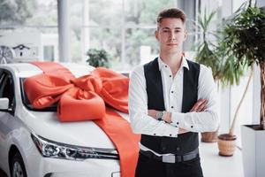 A handsome man is a buyer standing next to a new car at the dealer center and looking at the camera. photo