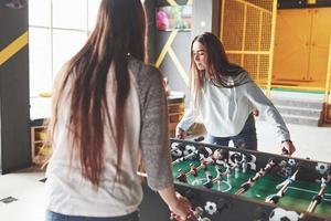 Two beautiful twin girls play table football and have fun. One of the sisters holds a toy ball in his hand and shows the tongue photo