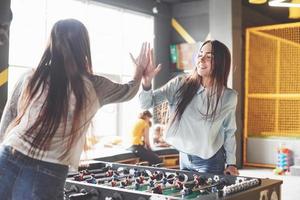 Two beautiful twin girls play table football and have fun. One of the sisters holds a toy ball in his hand and shows the tongue photo