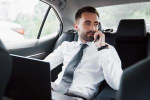 A young businessman working on laptop and talking on the phone while sitting in the car's back. Works in motion, appreciates its time