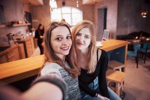 Two friends drinking coffee in a cafe, taking selfies with a smart phone and having fun making funny faces. Focus on the girl on the left photo