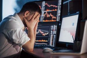 Stressful day at the office. Young businessman holding hands on his face while sitting at the desk in creative office. Stock Exchange Trading Forex Finance Graphic Concept photo