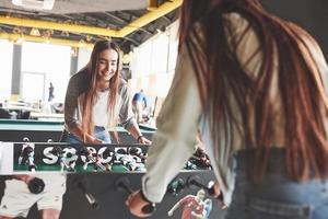 Two beautiful twin girls play table football and have fun. One of the sisters holds a toy ball in his hand and shows the tongue photo