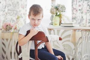 Small boy in blue trousers jeans is sitting on the wooden chair in the room photo