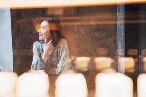 Mujer sonriente en la cafetería mediante teléfono móvil y mensajes de texto en las redes sociales, sentada sola