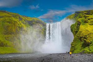 Powerful and famous Skogafoss waterfall with a lonely standing person in orange jacket, while hiking in Iceland, summer, scenic dramatic view at sunny day and blue sky. photo