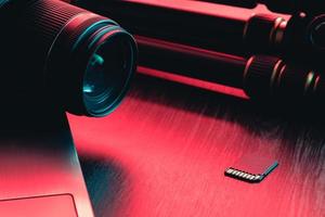 Camera lens, card, laptop and tripod on wood table. Desk workspace. Red and blue light photo